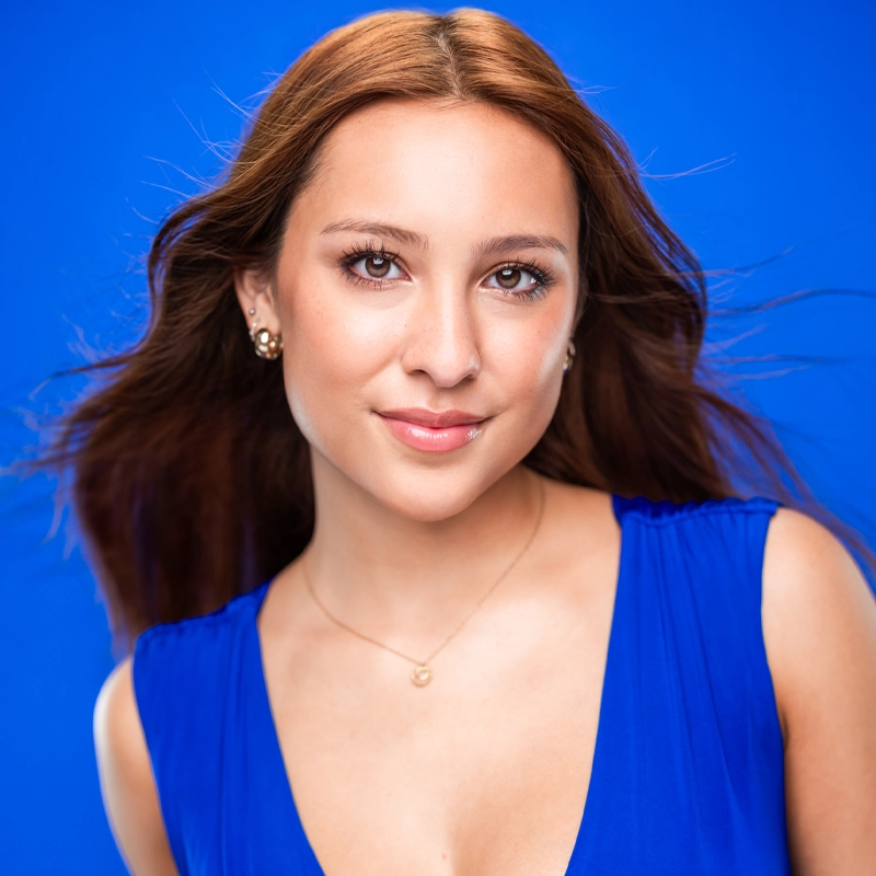 A young woman with long brown hair, wearing a sleeveless royal blue top and small gold jewelry, smiles softly at the camera. Her hair flows back, and the background is a solid, vibrant blue. This is Samantha Scott.
