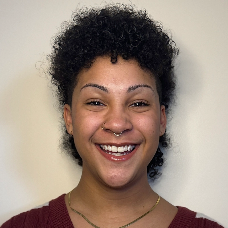 A person with short, curly dark hair and a septum piercing smiles in front of a plain light background. They wear a maroon sweater and a gold necklace. The photo is well-lit, showing clear facial features and a joyful expression—capturing Alexandra Zografos’s vibrant personality.