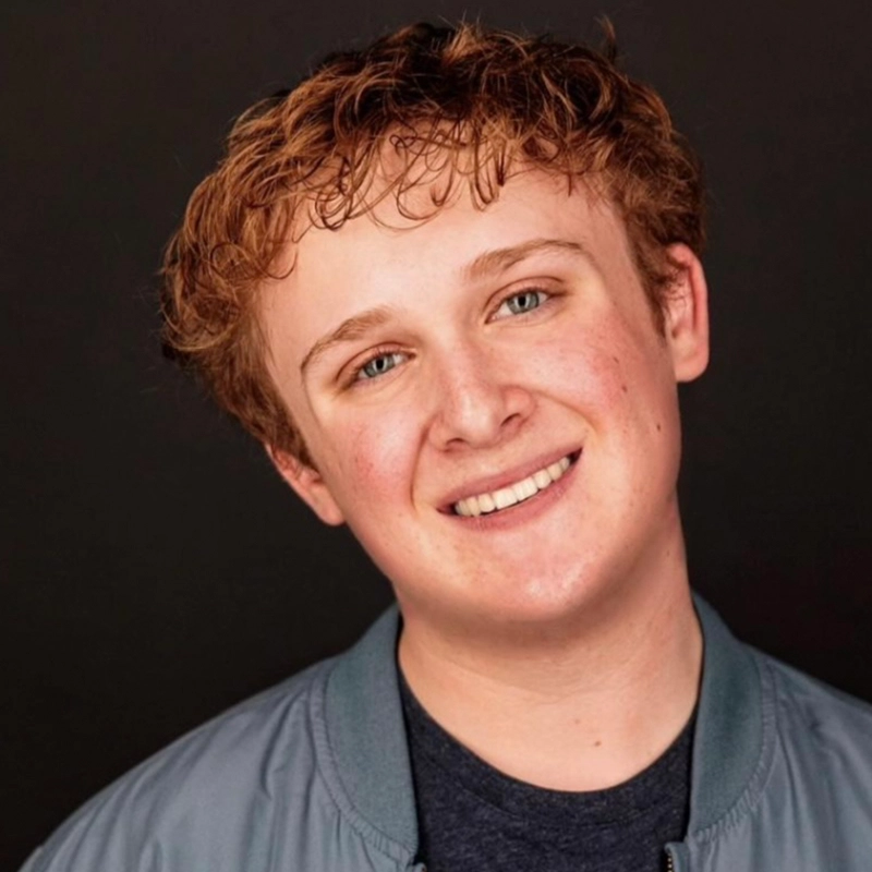 A young person with short, curly light brown hair smiles, looking directly at the camera. Megan Ludwig wears a blue-gray jacket over a dark shirt. The background is solid black. Their head is slightly tilted, and their skin is fair with a natural complexion.