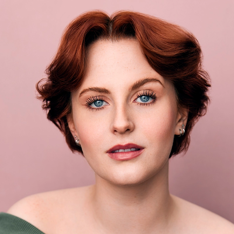 Gabrielle Farley, a person with short, wavy auburn hair, light skin, and blue eyes, looks at the camera. They wear subtle makeup, silver earrings, and an off-the-shoulder green top. The background is a soft pink color.
