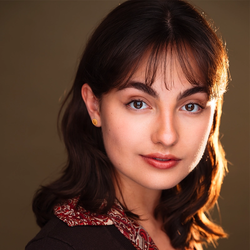 A young woman with medium-length brown hair and bangs gazes softly at the camera, reminiscent of a Gaël Mouton-Sady portrait. She has light skin, subtle makeup, and wears a maroon-patterned scarf over a dark top. The background is olive green and softly lit, highlighting her facial features.