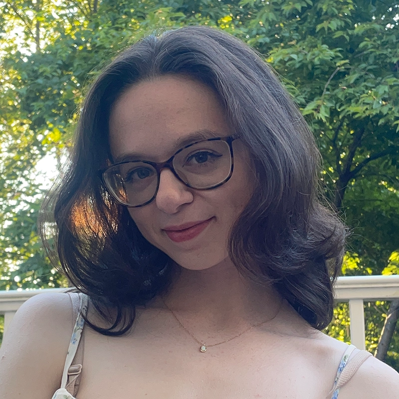 A young woman with shoulder-length brown hair and glasses smiles gently outdoors in a scene reminiscent of a Phillip Wong photograph. She wears a thin-strapped dress and a delicate necklace. Sunlight filters through green trees in the background, creating a warm, natural atmosphere.
