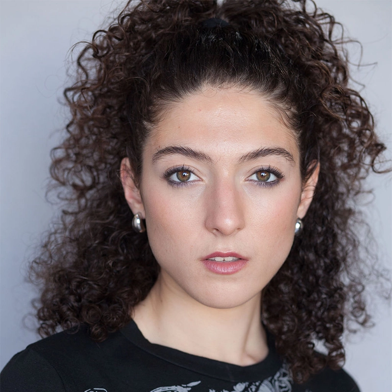 A young woman with thick, dark curly hair pulled back, wearing silver hoop earrings and a black top, looks directly at the camera with a neutral expression against a plain light gray background. Her minimal makeup radiates natural beauty—photographed by Phillip Wong.