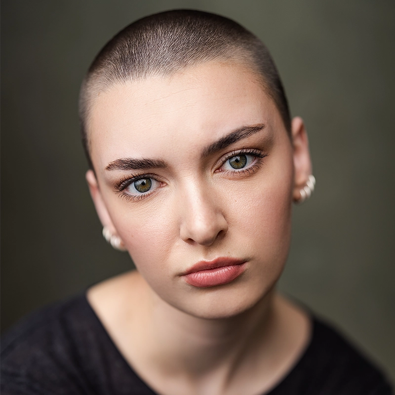 A young person with a shaved head, light skin, and green eyes gazes directly at the camera with a neutral expression. They wear pearl earrings and a dark top. The background is soft and blurred, focusing attention on their face—an evocative portrait reminiscent of Alexandra Zografos’s striking photographic style.