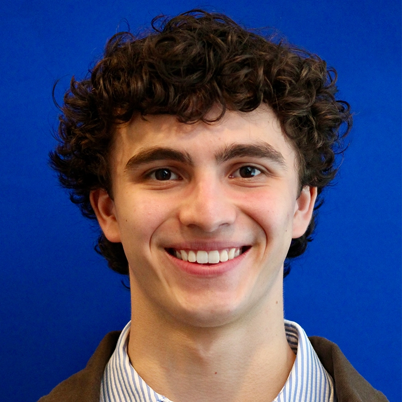 A young person with curly brown hair smiles, showing teeth. Casey Keeler wears a collared, blue-and-white striped shirt under a brown jacket. The background is solid bright blue.