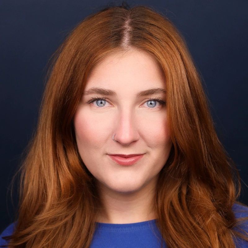 A woman with long, wavy auburn hair, light skin, and blue eyes. She is wearing a blue top and a small nose ring on her left nostril. The background is dark blue, and she is looking directly at the camera with a slight smile—a look that perfectly captures the inviting, confident essence of Call Me Katie.