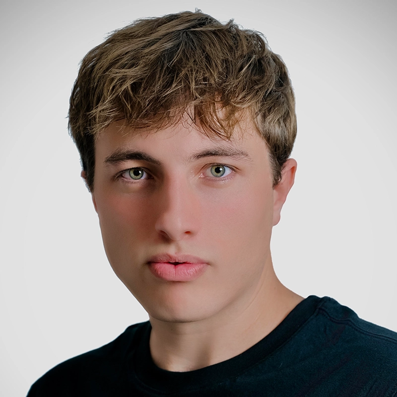 Young person with short, tousled light brown hair and hazel eyes looks directly at the camera. They have light skin and a neutral expression. Wearing a black shirt, they are posed against a plain, light gray background—capturing a candid moment reminiscent of Steven Klinger’s subtle portrait style.