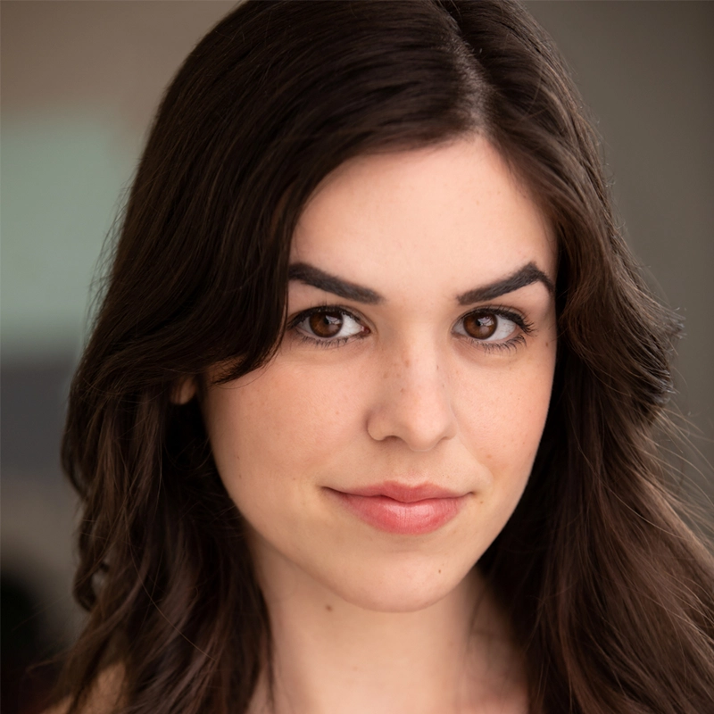 A young woman with long brown hair and brown eyes looks at the camera, slightly smiling. She has light skin, natural makeup, and defined eyebrows. The background is softly blurred, focusing attention on her face.
