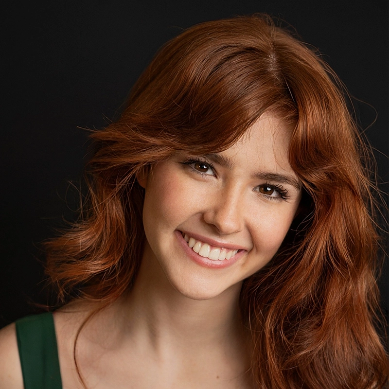 A young woman with long, wavy auburn hair smiles warmly. She has fair skin, light makeup, and is wearing a dark sleeveless top. The black background makes her hair and smile stand out, perfectly capturing Megan Ludwig’s radiant presence.