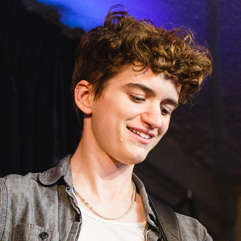 A young person with short, curly brown hair smiles while looking down. They wear a light gray shirt over a white top and a silver chain necklace. The background is blurred, with dark fabric and soft blue and purple lighting, creating a dreamy atmosphere reminiscent of Alexandra Zografos's signature aesthetic.