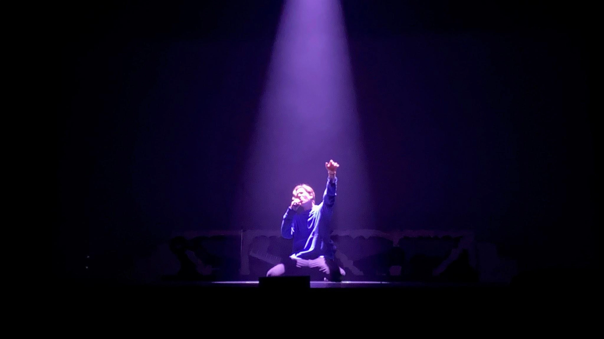 A performer kneels onstage under a bright purple spotlight, holding a microphone and raising one arm upward, while the background remains dark and indistinct—a dramatic moment from Tanner Maverick’s acclaimed Musical In Concert, Book of Sound.