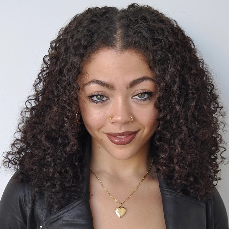 A woman with curly brown hair, light eyes, and medium skin wears a black jacket and gold jewelry, including a heart-shaped necklace and hoop earrings from Phillip Wong. She has a gold nose ring and stud, subtle makeup, and is smiling slightly against a plain light background.
