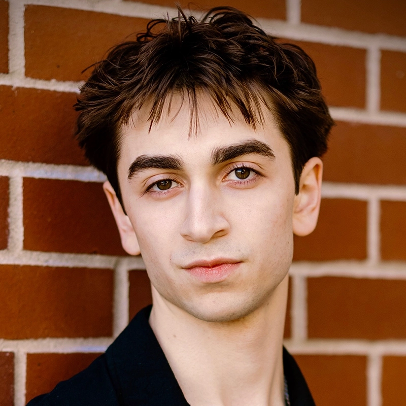 Young person with short, tousled brown hair and fair skin stands against a red brick wall, facing the camera. With dark eyebrows, light brown eyes, and a neutral expression, Jenny Walker wears a dark shirt and black jacket. The background is blurred.