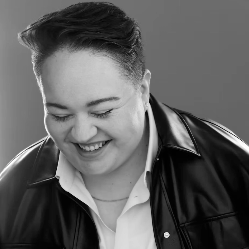Blayze Teicher, with short dark hair, smiles and looks down with eyes closed. Wearing a white collared shirt under a dark leather jacket, they radiate joy in this candid black-and-white photo set against a plain gray background.