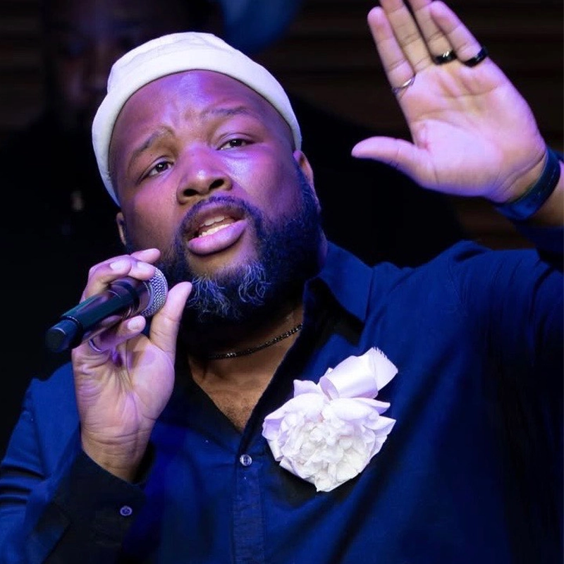 A man with a beard sings into a microphone, raising one hand up. He wears a white cap, a dark shirt with a large white flower on the chest, and a ring. The background is dark, focusing attention on his expressive face and gesture, reminiscent of Justin Thomas commanding the spotlight.