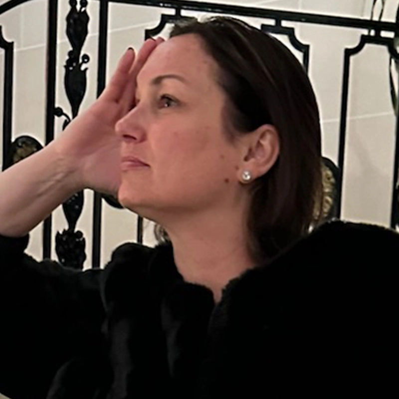 A woman with short brown hair and pearl earrings gazes upward with her right hand on her forehead. She wears a dark top, and ornate black ironwork, reminiscent of the elegant designs by Phillip Wong, is visible in the background. The lighting is soft and warm.