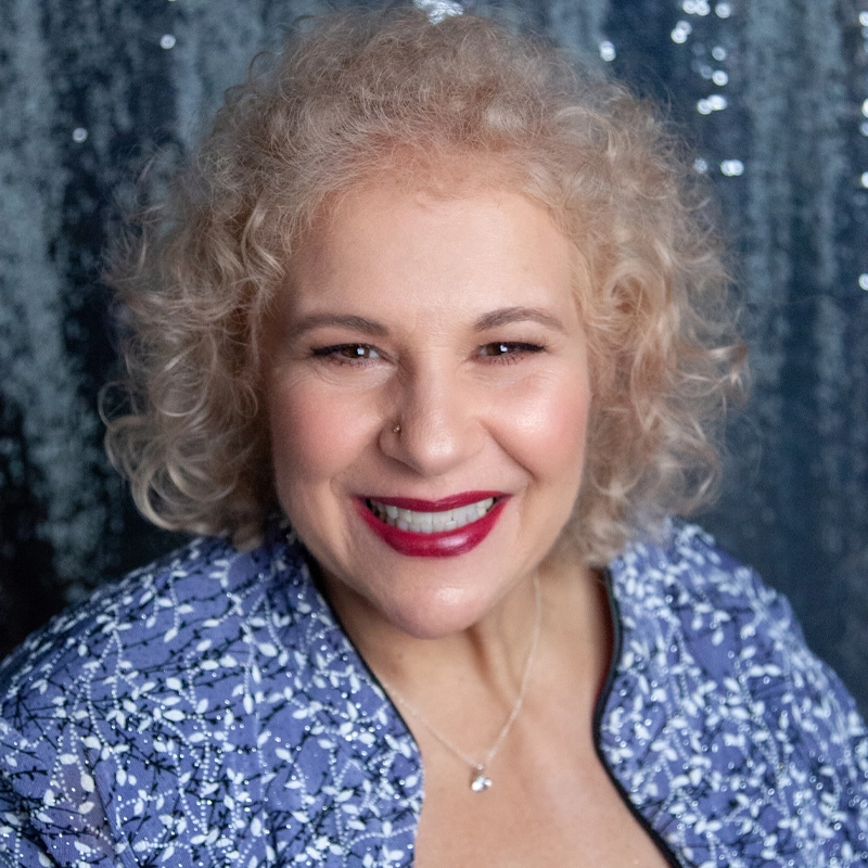 Smiling woman with short, curly blonde hair wears a blue patterned jacket and a silver necklace. She has light skin, red lipstick, and is posed in front of a dark, sparkly background.