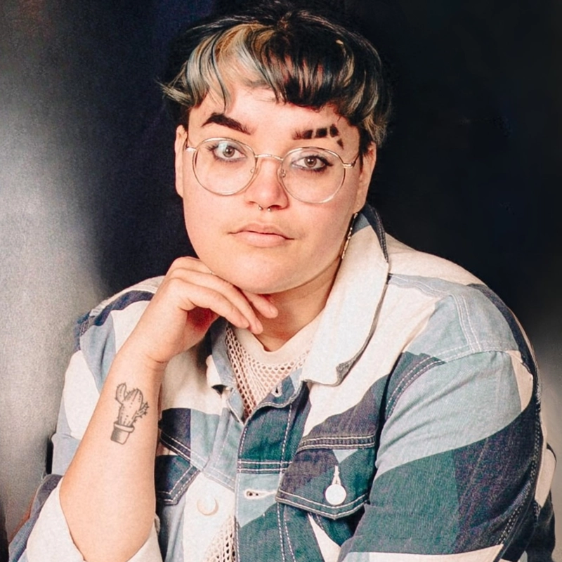 A person with short, curly hair dyed black and light blue, wearing round glasses and a patchwork denim jacket. They rest their chin on their hand, showing a cactus tattoo on their forearm, and have bold eyebrows with shaved lines. The background is dark.