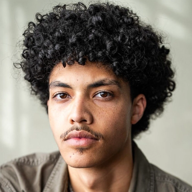 A person with light brown skin, short curly black hair, and a thin mustache looks directly at the camera with a neutral expression, wearing a green jacket. The background is softly blurred and light gray, creating a subtle focus on Asher Alcantara.