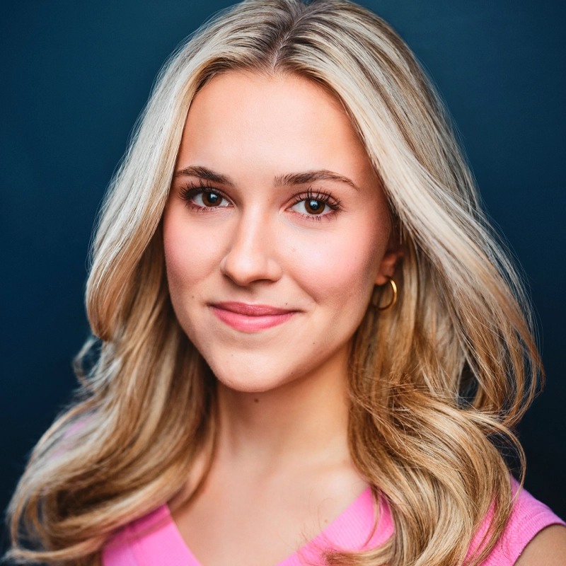 A young woman with long, wavy blonde hair smiles softly. She has light skin, brown eyes, and wears a pink top with gold hoop earrings. The background is a solid dark blue. Her hair is parted in the middle and falls over her shoulders, capturing the approachable charm of Taylor Truckenbrod.