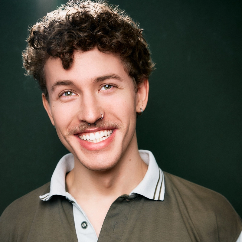 Brennan Trudel, a young person with curly brown hair, a mustache, and a nose ring, smiles warmly. They wear an olive green and light gray collared shirt, set against a dark green background with soft lighting highlighting their face.