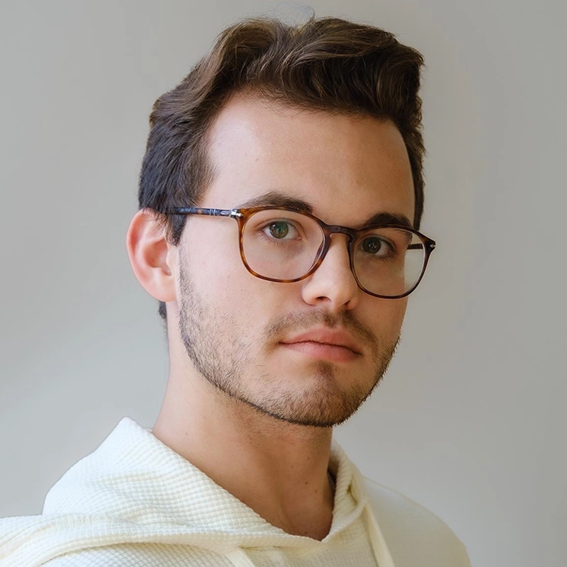 Young man with short brown hair, light beard, and wearing round eyeglasses, faces forward with a neutral expression. He is dressed in a light-colored hoodie and stands against a plain, light grey background. The lighting is soft and even. This portrait features Nic Sylvester.