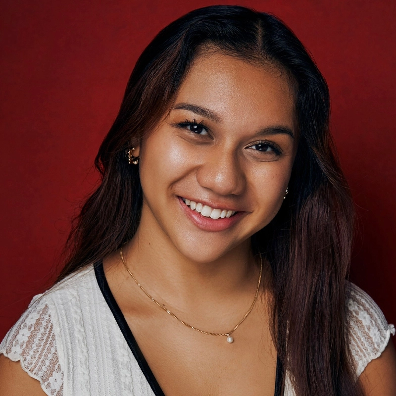A young woman with long, brown hair smiles warmly at the camera. She wears a white, lace-trimmed top and a delicate gold necklace with a small pearl. The background is solid red. Her makeup is subtle and her style is natural and elegant, perfectly capturing Marielle Utayde’s timeless charm.