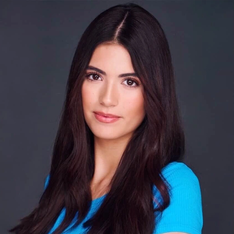 A young woman with long, dark brown hair poses against a plain dark background. She has fair skin, natural makeup, and is wearing a bright blue shirt. Lauren Velasco looks at the camera with a neutral, soft expression.