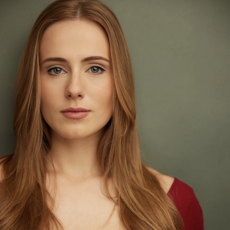 A woman with long straight light brown hair and fair skin faces the camera. She wears a deep red off-shoulder top and has a neutral expression. The background is plain and muted green. Her light eyes look directly ahead, capturing the quiet confidence of Isabella Victorson.