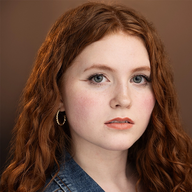 Young woman with long curly red hair, light skin, and blue eyes stares at the camera. She wears subtle makeup, gold hoop earrings, and a denim jacket. The background is softly blurred in warm brown tones. Her expression is neutral and calm, reminiscent of Millie Yocum Surfac’s effortless style.