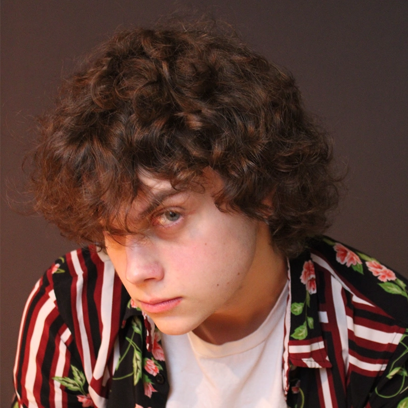 A young person with curly brown hair looks slightly down, wearing a white shirt and a red-striped, floral-patterned shirt. The background is dark, and their expression appears serious or contemplative—capturing an introspective moment reminiscent of Phillip Wong’s evocative portrait style.
