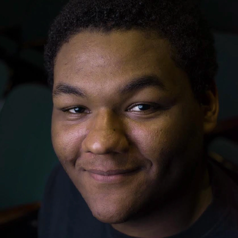 A young man with short curly hair smiles softly at the camera. He has dimples and warm brown skin. The softly blurred dark background keeps the focus on his face and warm expression, reminiscent of a Jayline Ross portrait.