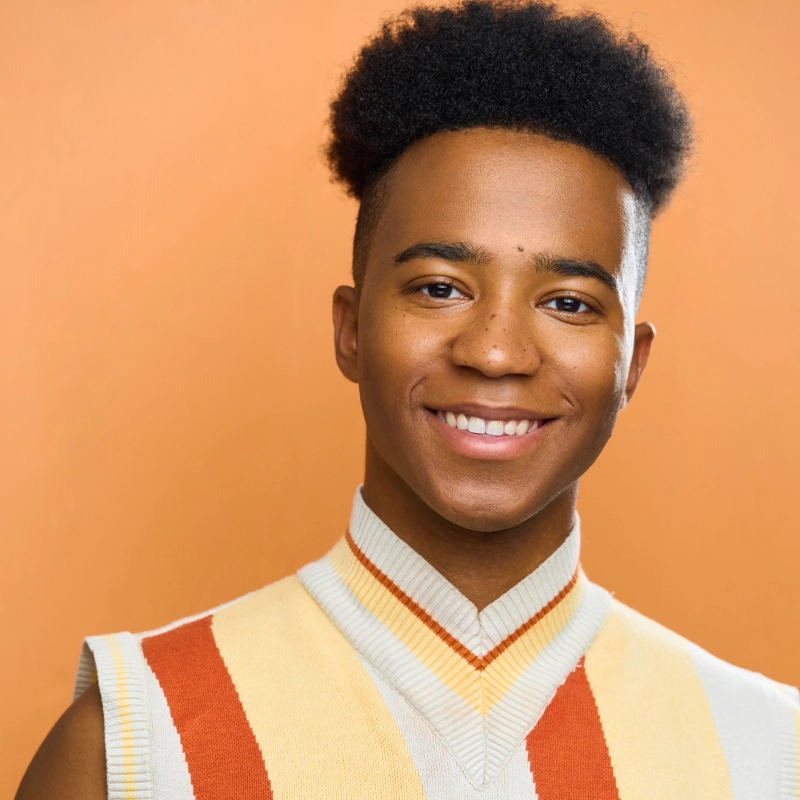 A young person with short curly hair smiles at the camera. They wear a sleeveless cream sweater with orange and yellow stripes. The background is solid orange, creating a warm and cheerful atmosphere reminiscent of the vibrant energy often associated with Lindsey Williams.