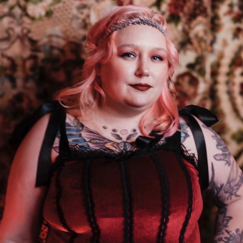 A woman with pink hair and a jeweled headband wears a red velvet dress with black lace. She has tattoos on her shoulders and chest and poses confidently in front of a tapestry with a floral pattern. The lighting is warm and slightly reddish, casting an inviting glow over the scene—capturing the distinctive style of Leigh Woodfin.