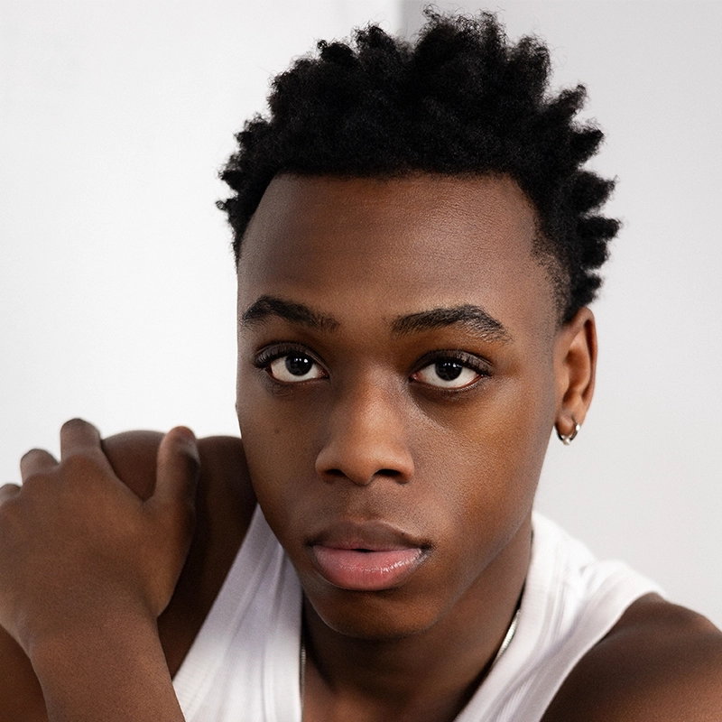 A young Black man with short, natural textured hair wears a white tank top and small hoop earrings. He looks directly at the camera with a neutral expression, resting one arm on his shoulder against a plain light background. The portrait captures the understated confidence of Gaël Mouton-Sady.