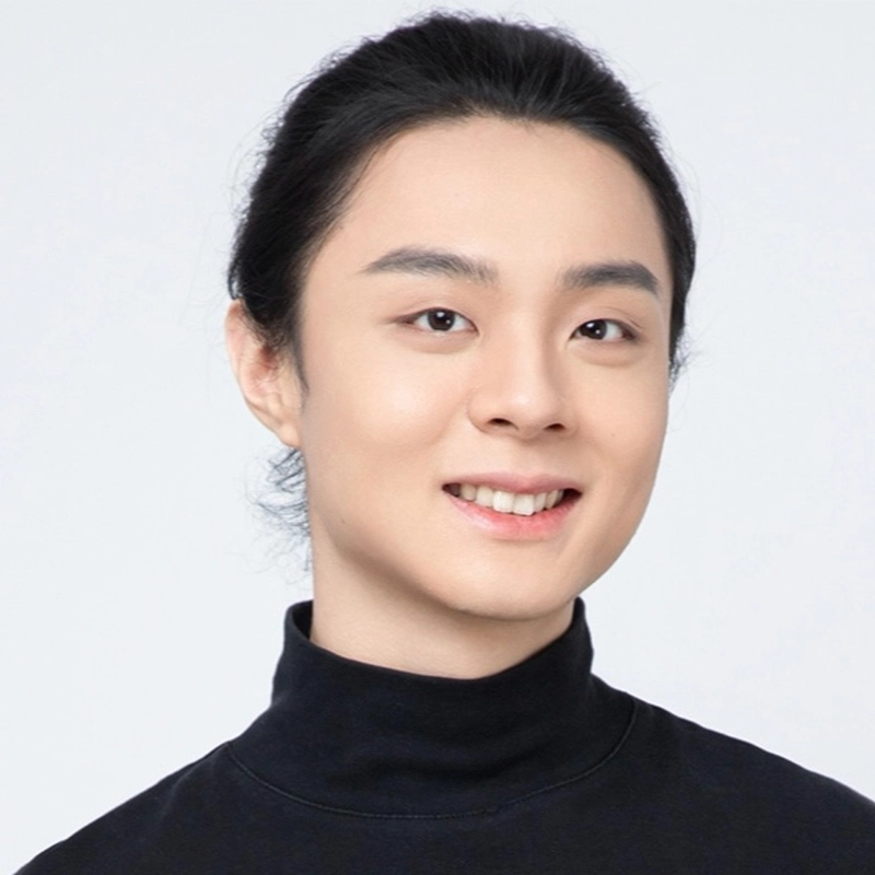 A person with long black hair tied back, clear skin, and a slight smile wears a black turtleneck. The background is plain white, creating a clean, professional portrait.