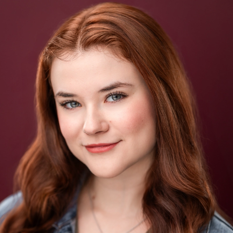 Young woman, Jayline Ross, with long, wavy auburn hair, fair skin, and blue eyes smiles softly. She wears natural makeup and a denim jacket. The solid maroon background adds warmth and professionalism to the portrait.