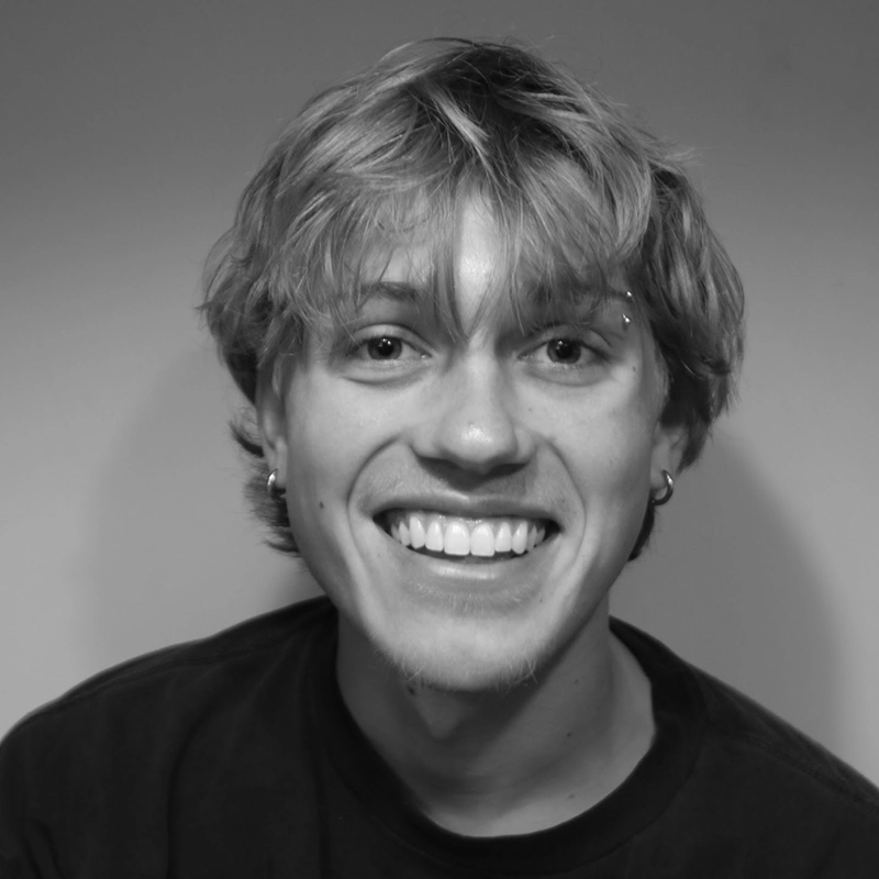 A young person with wavy, light hair smiles widely at the camera. They wear earrings, a dark t-shirt, and have an eyebrow piercing. The background is plain and the photo is in black and white, capturing Gaël Mouton-Sady's effortless style and charismatic presence.