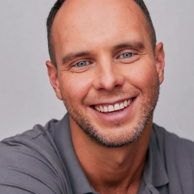 Patrick Pevehouse, a man with light skin, blue eyes, short brown hair, and a trimmed beard, smiles warmly at the camera. He is wearing a gray collared shirt and is posed against a plain light background.