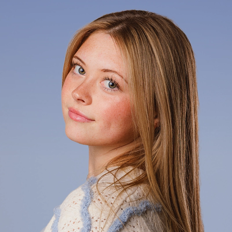 Teen girl Jenny Walker with long straight blonde hair and blue eyes, wearing a cream sweater with blue trim, looks over her shoulder and smiles softly. Her skin is fair with light freckles. The background is solid light blue, creating a calm and serene atmosphere.