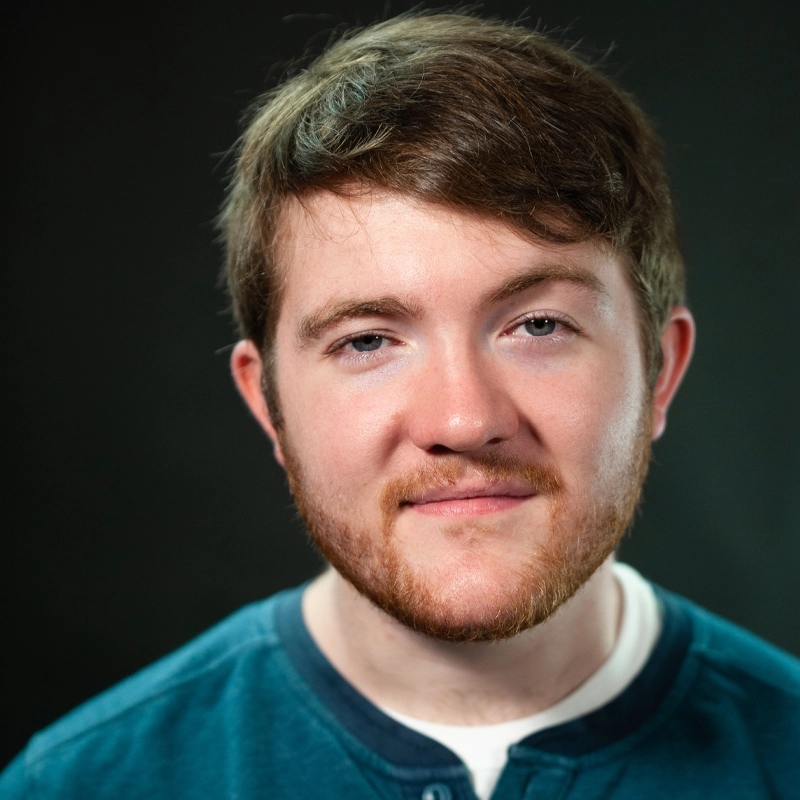 Young man with short brown hair and a light brown beard smiles softly at the camera. He wears a teal shirt over a white undershirt. The background is dark and plain, putting focus on his face. The lighting is bright and even.