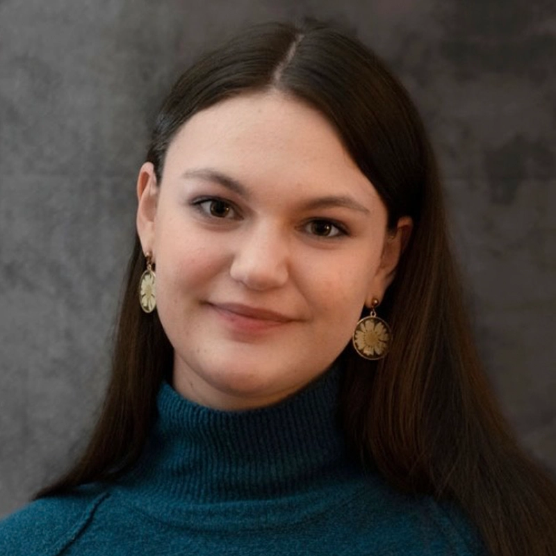 A young woman with straight brown hair wears a teal turtleneck sweater and large circular gold earrings. Abby Gorsuch smiles softly at the camera. The background is a blurred gray pattern.