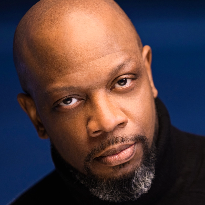 A bald, middle-aged Black man with a short gray beard and mustache, wearing a black turtleneck sweater, gazes directly at the camera with a serious expression against a blue background—this is Melvin Abston.
