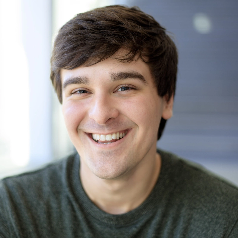 A young man with short brown hair smiles warmly at the camera. He is wearing a dark green sweater. The background is softly blurred with blue and white tones, suggesting a bright indoor setting. The lighting is natural, highlighting his friendly expression—an inviting portrait of Luis Acevedo.