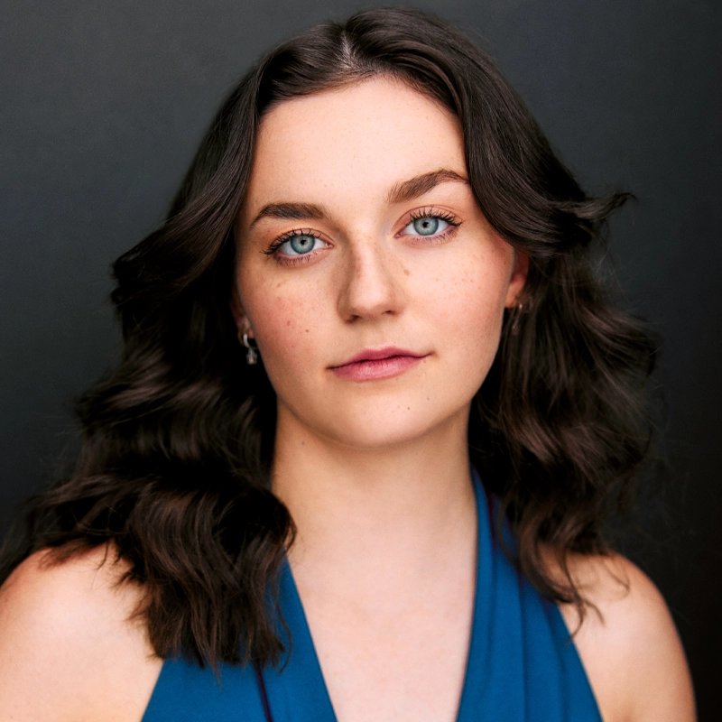 A young woman with wavy dark brown hair and blue eyes faces the camera against a dark background. She has fair skin, light makeup, and is wearing a sleeveless blue top. With a calm, neutral expression, Lyndsey Adkisson’s small earrings are visible.
