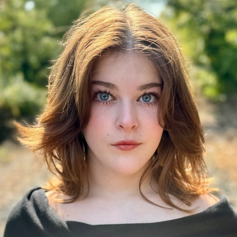 Young woman with shoulder-length light brown hair, blue eyes, and fair skin stands outdoors. Celeste Zwingel wears a black top and looks directly at the camera, with a soft natural background of green foliage and sunlight.