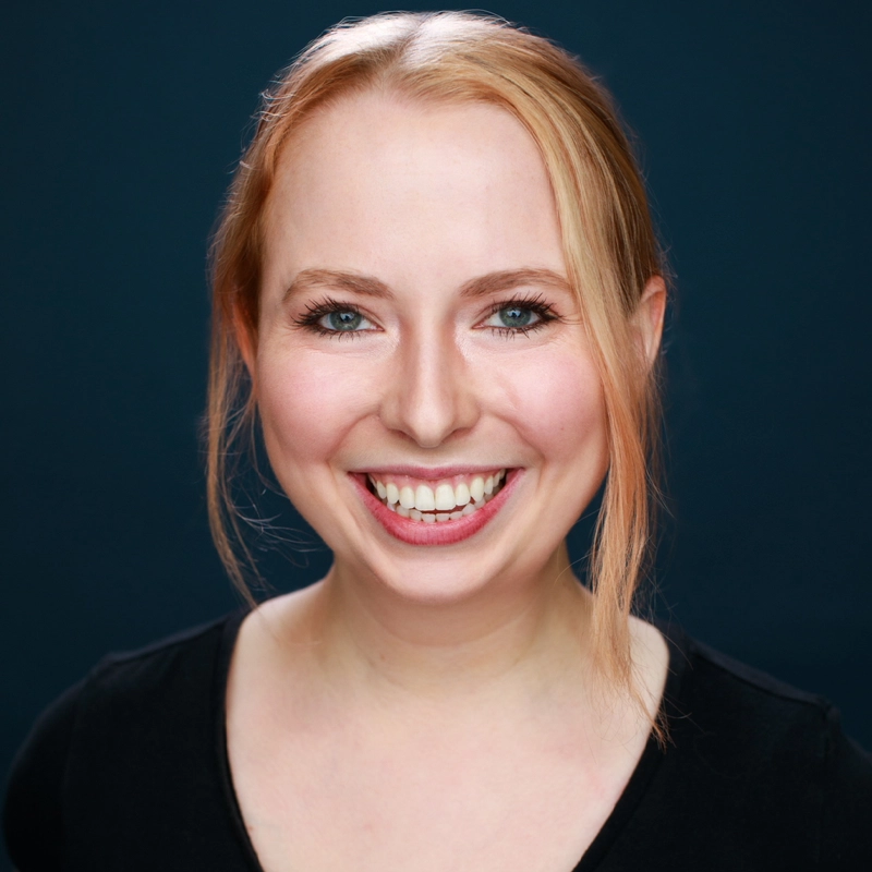 A young woman with fair skin and strawberry blonde hair smiles widely, showing her teeth. She has blue eyes and is wearing a black top. Zayda Albert’s hair is parted slightly off-center and pulled back with a few loose strands framing her face. The background is a deep, sage-tinged dark blue.