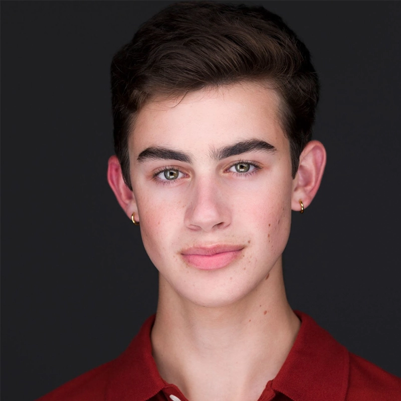 A young person with short brown hair, light eyes, and fair skin looks at the camera. They wear small gold hoop earrings and a red collared shirt. The plain, dark background highlights their face in natural light, capturing the quiet confidence reminiscent of Celeste Zwingel.