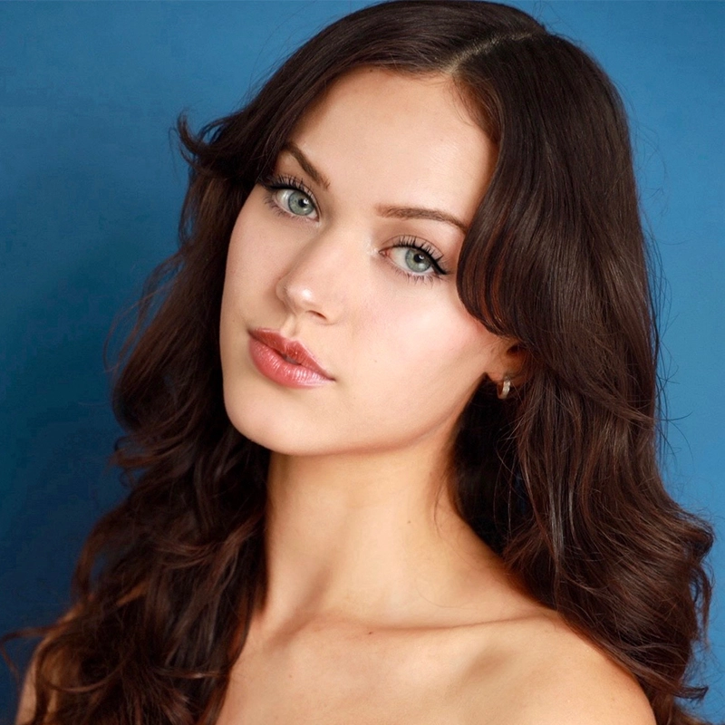 A young woman with long, wavy brown hair poses against a blue background in an Aaron Ballentine-inspired portrait. She has light eyes, clear fair skin, and natural makeup with subtle lip gloss. She wears small earrings and no visible clothing straps, giving the image a slightly formal, portrait feel.