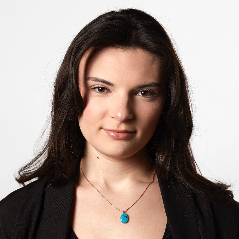 A woman with long brown hair wearing a black top and jacket, along with a turquoise pendant necklace, looks at the camera with a neutral expression. The style is reminiscent of Willow Wakar against a plain white background.